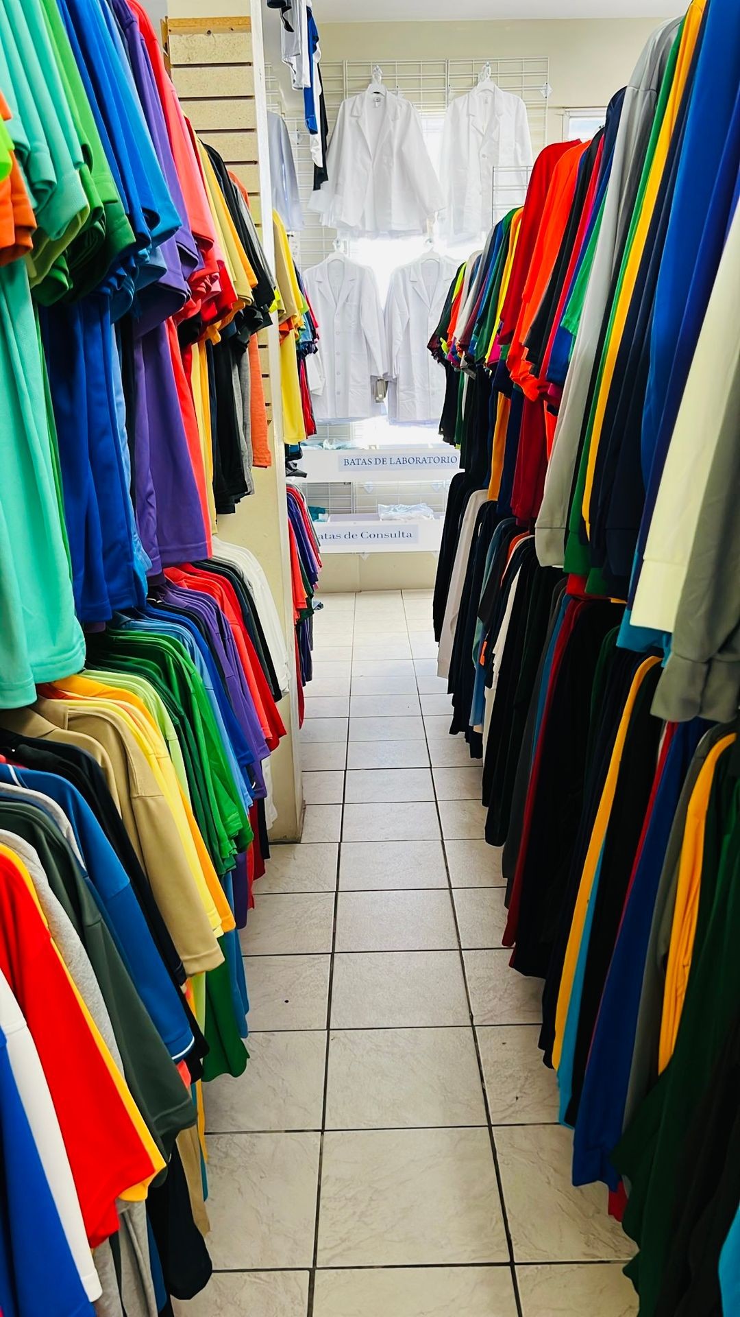 Narrow aisle in clothing store lined with colorful shirts and lab coats hanging on racks.